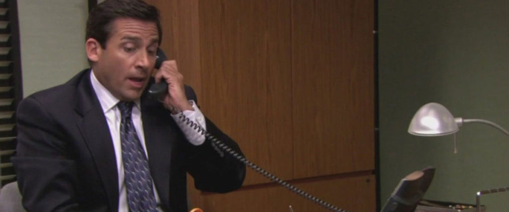 A man in a dark suit and patterned tie is sitting at a desk, holding a phone to his ear, speaking with a surprised expression. On the desk, which has a lamp and some office supplies, lies an open pitch deck. In the background, you can see a wooden cabinet and part of a gray wall.