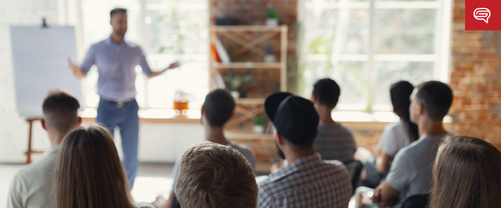 A group of people sitting in a classroom-like setting attentively listen to a man who is standing and gesturing in front of a flipchart, harnessing marketing principles. The room has a brick wall, large windows, and a shelving unit in the background.