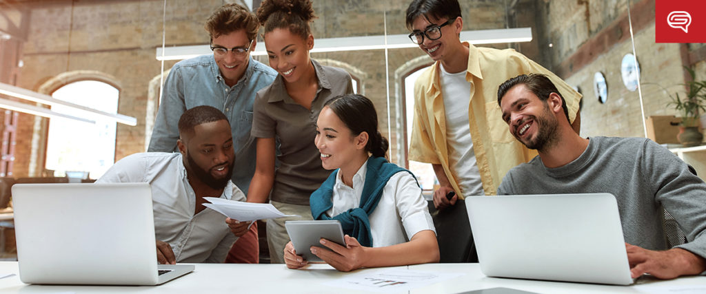 A diverse group of six people in a modern office collaborates enthusiastically around a table with laptops and documents. They seem to be discussing something on a tablet held by one of the participants, all smiling and engaged, emerging strong from recent crises.