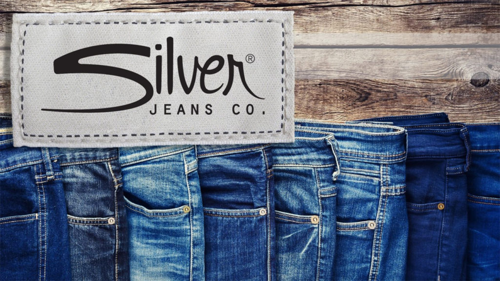 A pitch deck slide with a fabric tag reading "Silver Jeans Co." is stitched onto a wooden background. Below the tag, several pairs of Silver Jeans in different shades of blue are lined up side by side, displaying their back pockets and waistbands.
