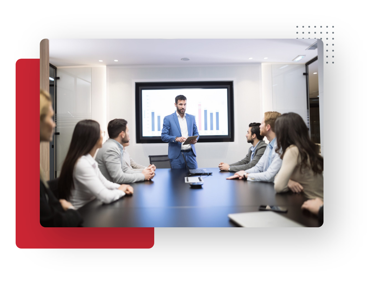 A man in a blue suit stands at the head of a conference table, presenting small business bar charts to a group of six people. The team listens attentively in a modern office setting as data is displayed on the large screen behind him.